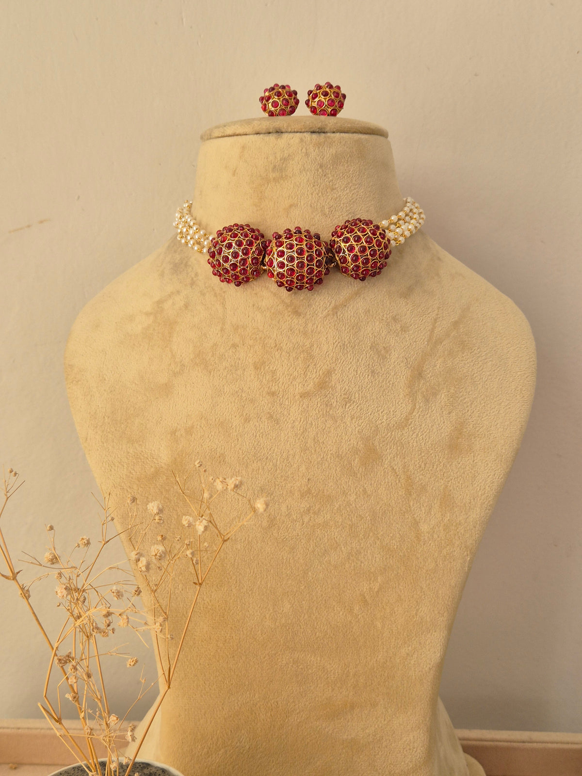 Aakriti Necklace Set featuring gold-toned spherical elements encrusted with red stones, paired with multi-strand seed pearl accents and matching round stud earrings, displayed on a beige jewelry mannequin by Naitri.in