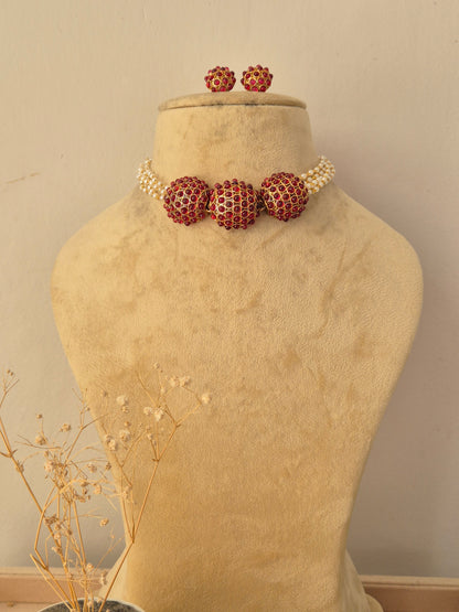Aakriti Necklace Set featuring gold-toned spherical elements encrusted with red stones, paired with multi-strand seed pearl accents and matching round stud earrings, displayed on a beige jewelry mannequin by Naitri.in