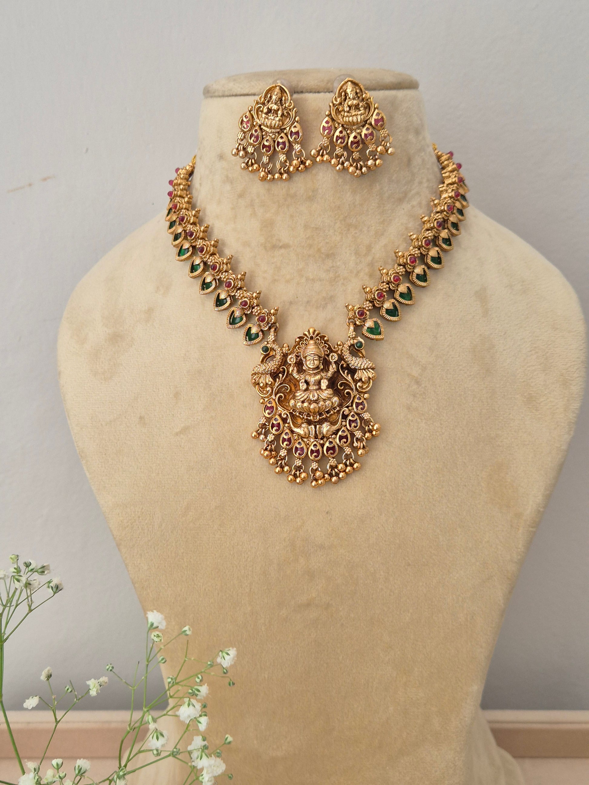 A gold-plated temple jewelry necklace featuring a detailed Goddess Lakshmi pendant with green and pink stone embellishments, with matching hanging jhumka earrings by naitri.in