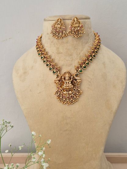 A gold-plated temple jewelry necklace featuring a detailed Goddess Lakshmi pendant with green and pink stone embellishments, with matching hanging jhumka earrings by naitri.in
