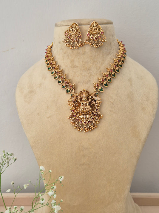 A gold-plated temple jewelry necklace featuring a detailed Goddess Lakshmi pendant with green and pink stone embellishments, with matching hanging jhumka earrings by naitri.in