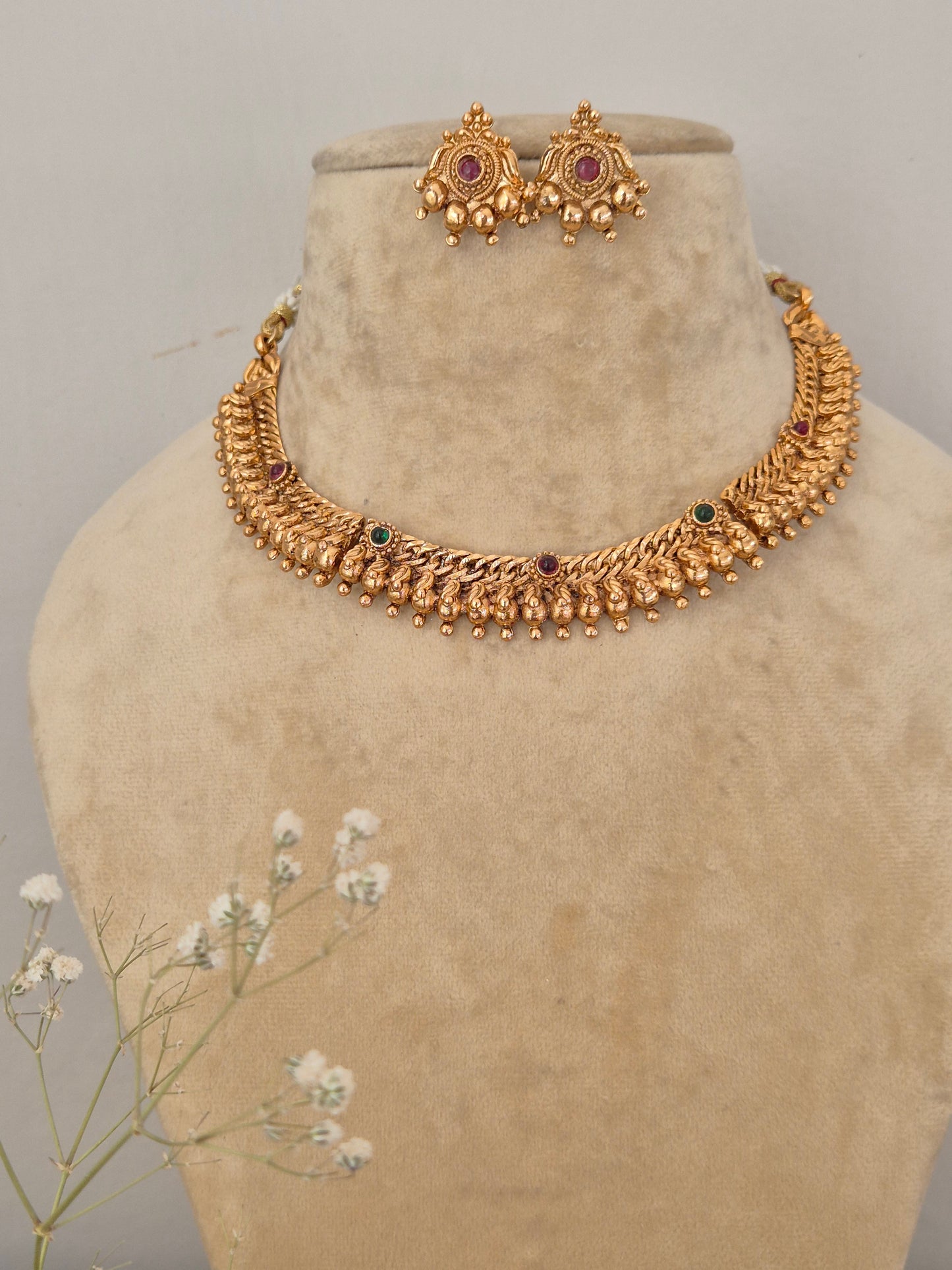 Gold-polished textured ethnic choker with red and green stone embellishments, dangling gold beads, and coordinating festive earrings by naitri.in