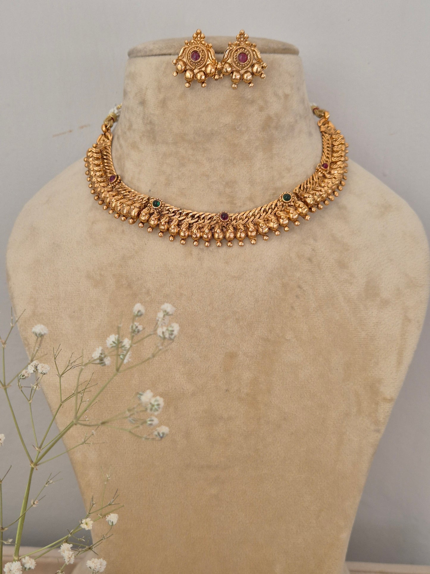 Handcrafted gold-plated choker necklace featuring intricate leaf-shaped textured metalwork, ruby-red and emerald-green stone accents, and a dangling gold bead fringe, with matching round stud earrings by naitri.in