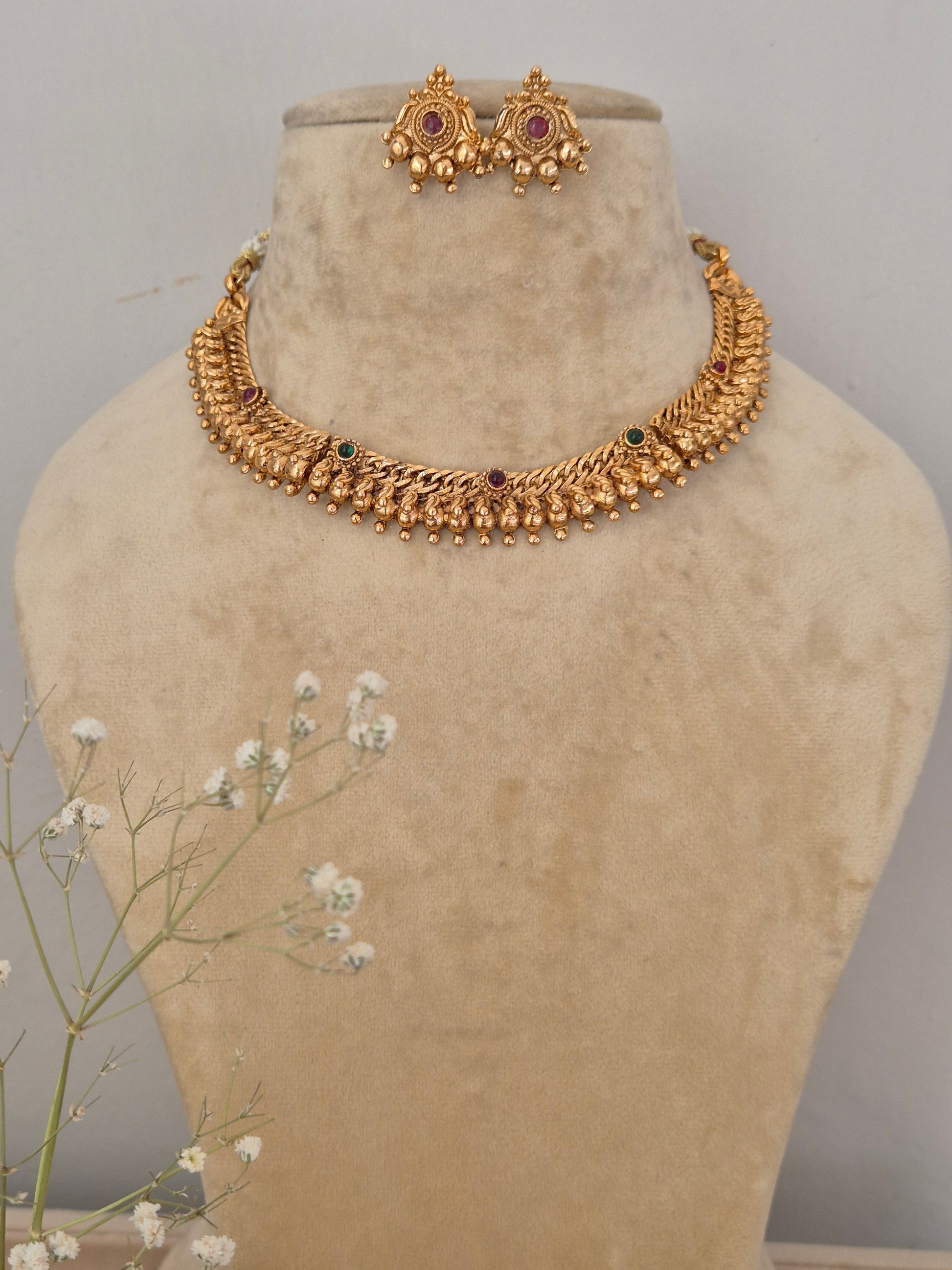 Handcrafted gold-plated choker necklace featuring intricate leaf-shaped textured metalwork, ruby-red and emerald-green stone accents, and a dangling gold bead fringe, with matching round stud earrings by naitri.in