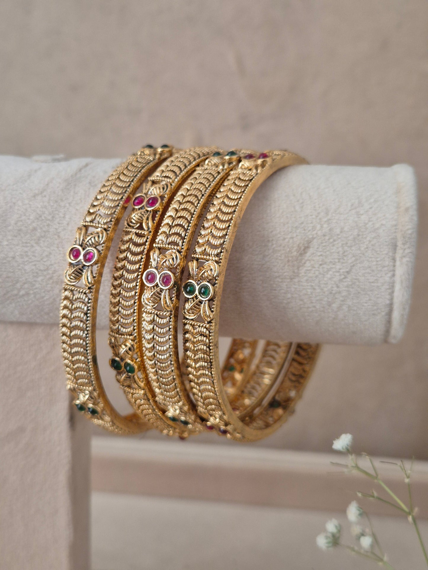 A stack of four Naitri Ahilya gold bangles showcasing detailed ethnic craftsmanship, embedded with vibrant pinkish-red and green accent stones, by naitri.in