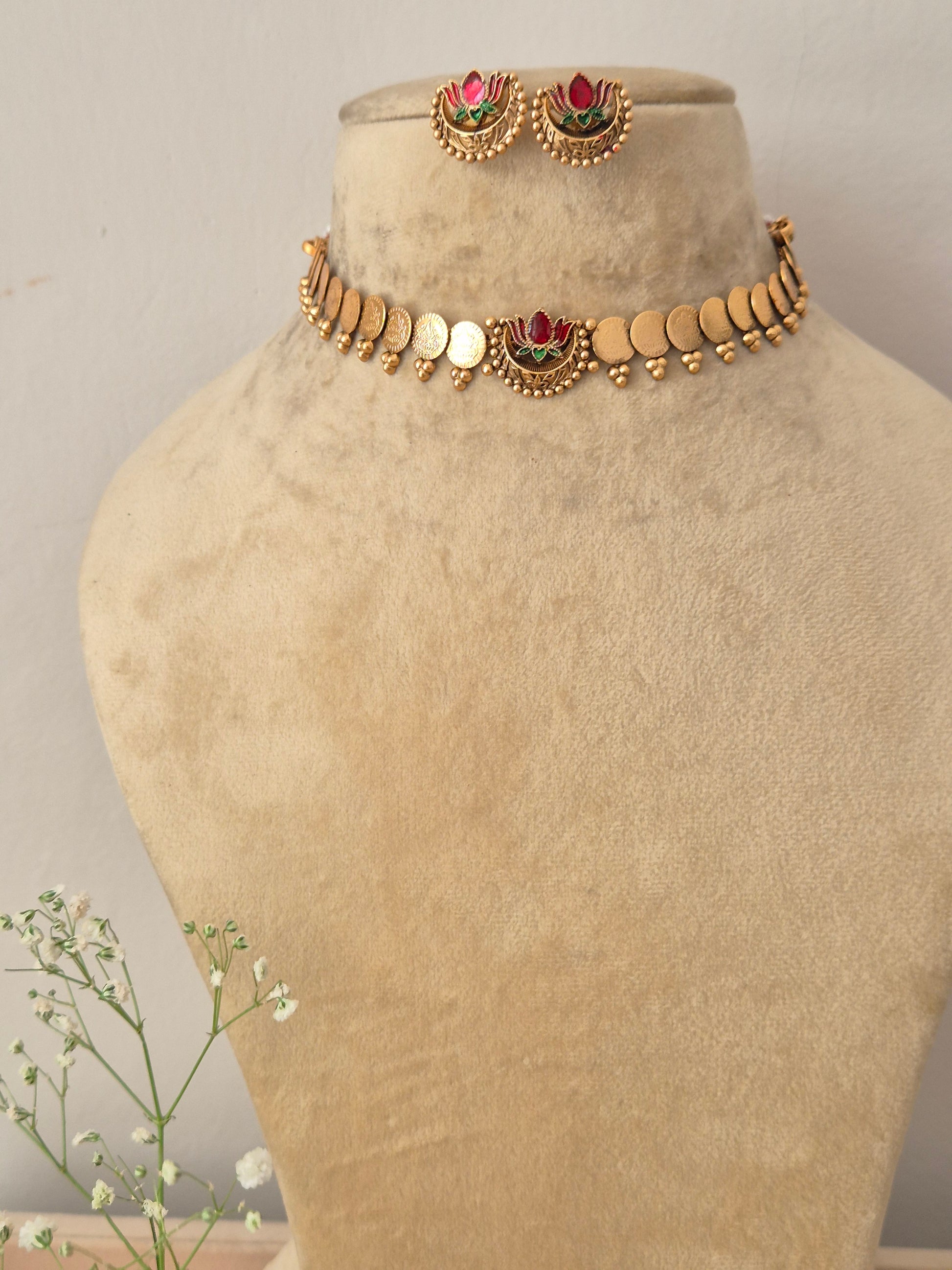 A gold-plated traditional choker necklace featuring small coin charms and a central pink and green lotus motif, displayed on a beige mannequin bust with matching lotus stud earrings by naitri.in