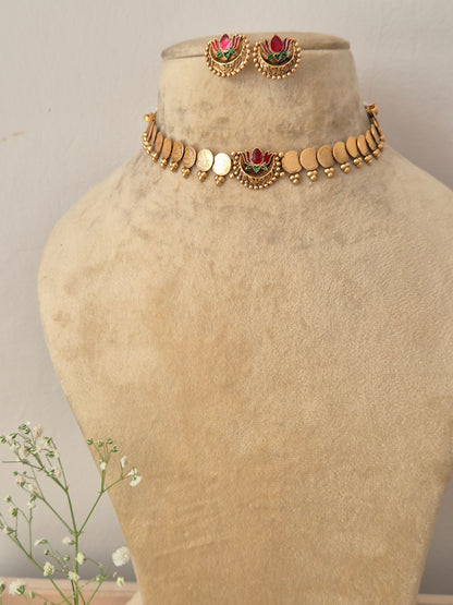 A gold-plated traditional choker necklace featuring small coin charms and a central pink and green lotus motif, displayed on a beige mannequin bust with matching lotus stud earrings by naitri.in