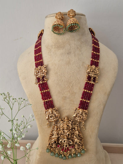 A traditional gold-plated temple jewelry necklace featuring multi-strand red beads and a large, intricate Goddess pendant with green accents, with matching jhumka earrings by naitri.in