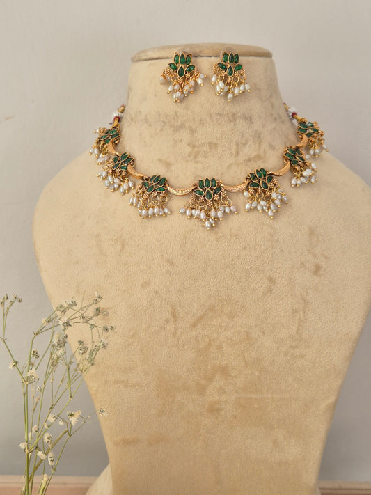 Gold-toned traditional Indian choker necklace and matching dangle earrings set features a series of delicate gold-toned floral motifs inset with vibrant green gemstones and a lower fringe of hanging small white pearls, by naitri.in