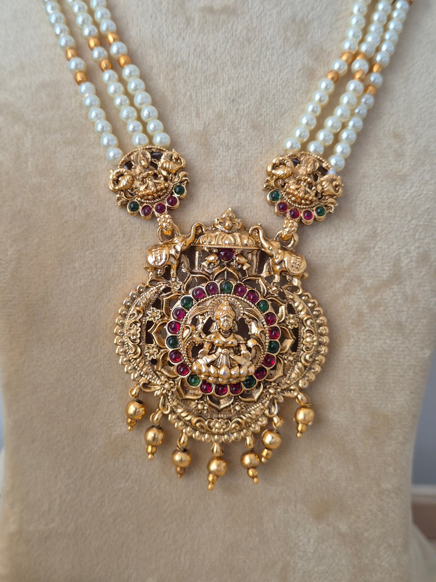 Three-layered white pearl chain necklace featuring a heavily detailed gold-toned Lakshmi pendant with hanging bead drops, paired with matching ornate bell-shaped earrings by naitri.in