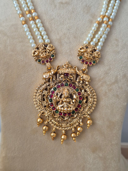 Three-layered white pearl chain necklace featuring a heavily detailed gold-toned Lakshmi pendant with hanging bead drops, paired with matching ornate bell-shaped earrings by naitri.in