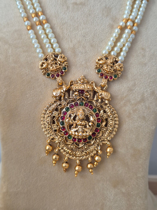Three-layered white pearl chain necklace featuring a heavily detailed gold-toned Lakshmi pendant with hanging bead drops, paired with matching ornate bell-shaped earrings by naitri.in