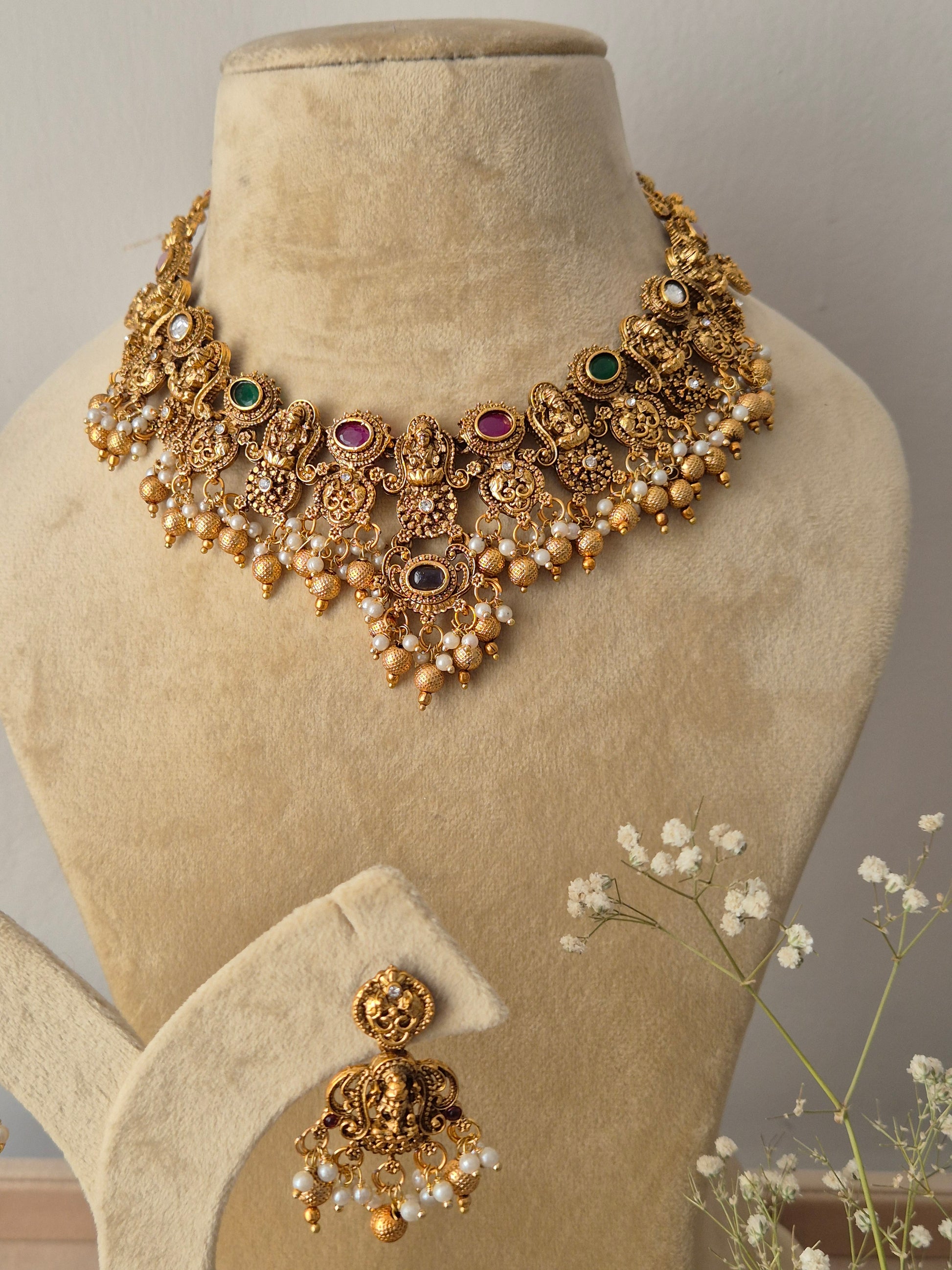 Handcrafted gold temple jewelry choker necklace and matching jhumka earrings with red and green stones Gold-polished temple choker with red and green stone embellishments, dangling gold beads, and coordinating festive jhumkas by naitri.in