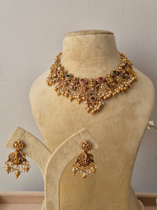 Handcrafted antique gold-plated temple jewelry choker necklace featuring a central deity motif, alternating ruby-red and emerald-green stones, and gold bead fringes, with matching jhumka drop earrings by naitri.in