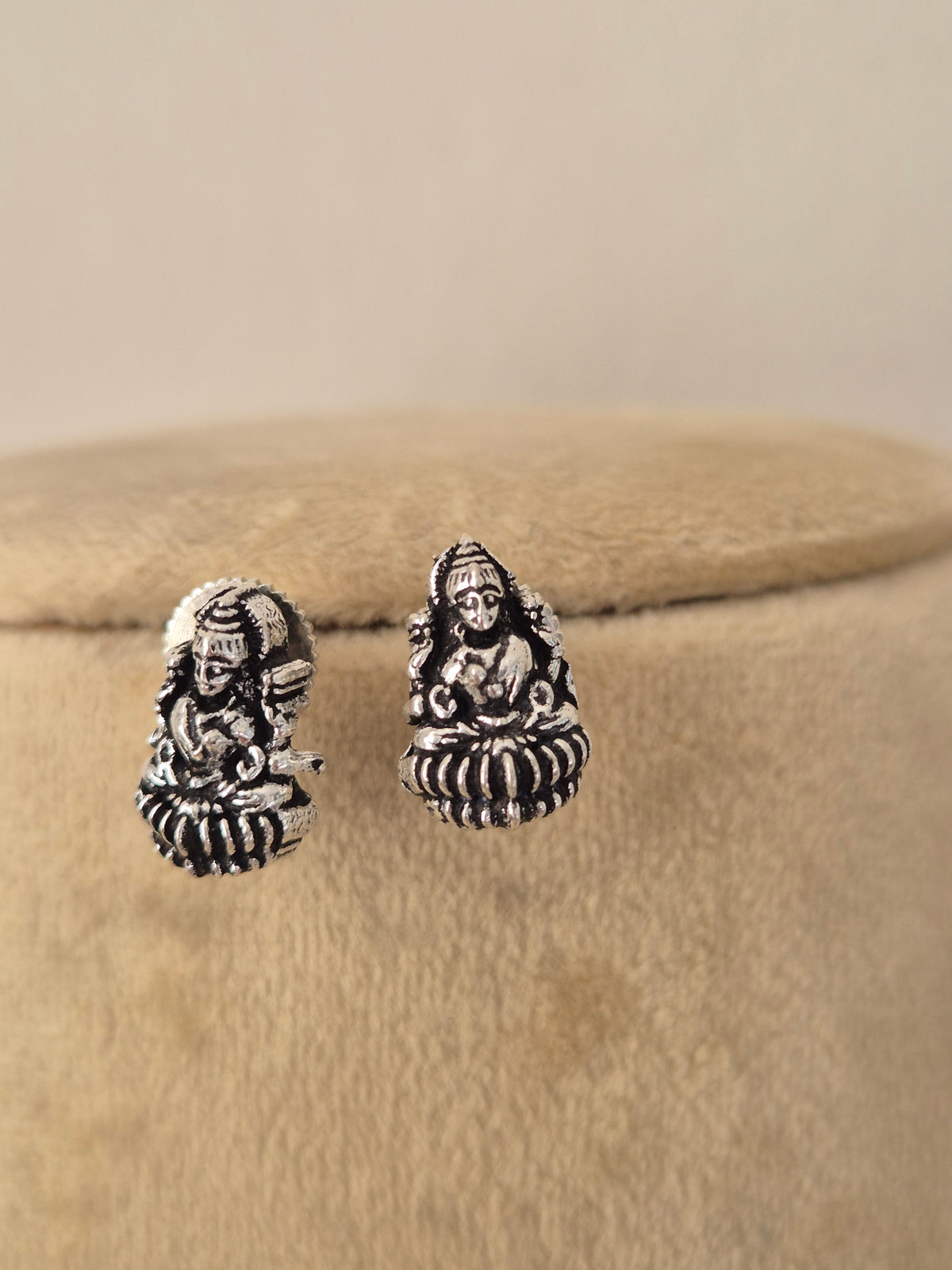 A pair of silver-toned stud earrings featuring detailed Hindu deity motifs, displayed on a beige velvet jewelry stand against a neutral beige background. by naitri.in