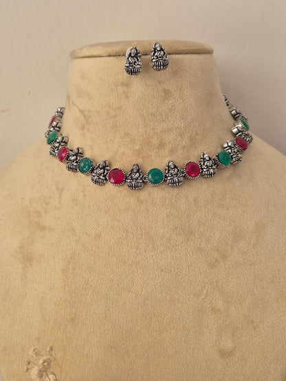 A silver-toned jewelry set , featuring a necklace and matching earrings with small seated deity motifs and round red and green gemstone accents. by naitri.in