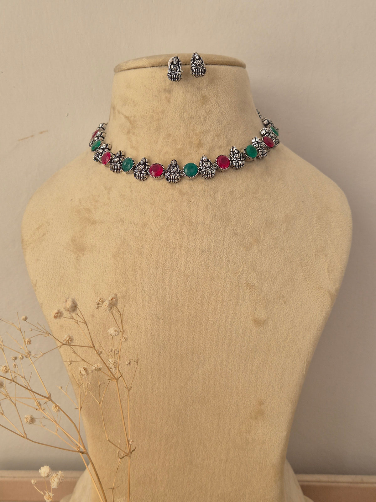 A silver-toned jewelry set  featuring a choker necklace and matching earrings with small Buddha figurines and round red and green gemstone accents by naitri.in