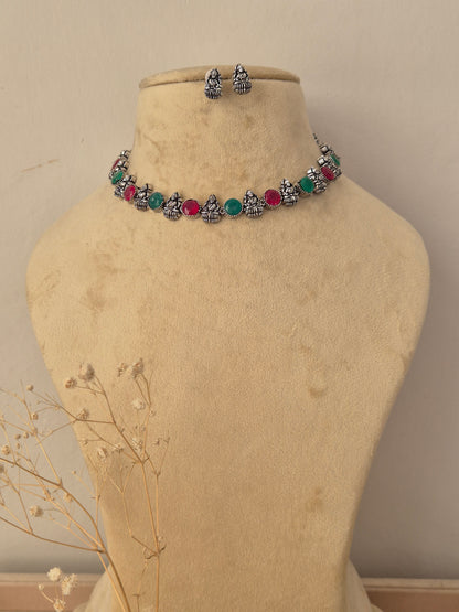 A silver-toned jewelry set  featuring a choker necklace and matching earrings with small Buddha figurines and round red and green gemstone accents by naitri.in