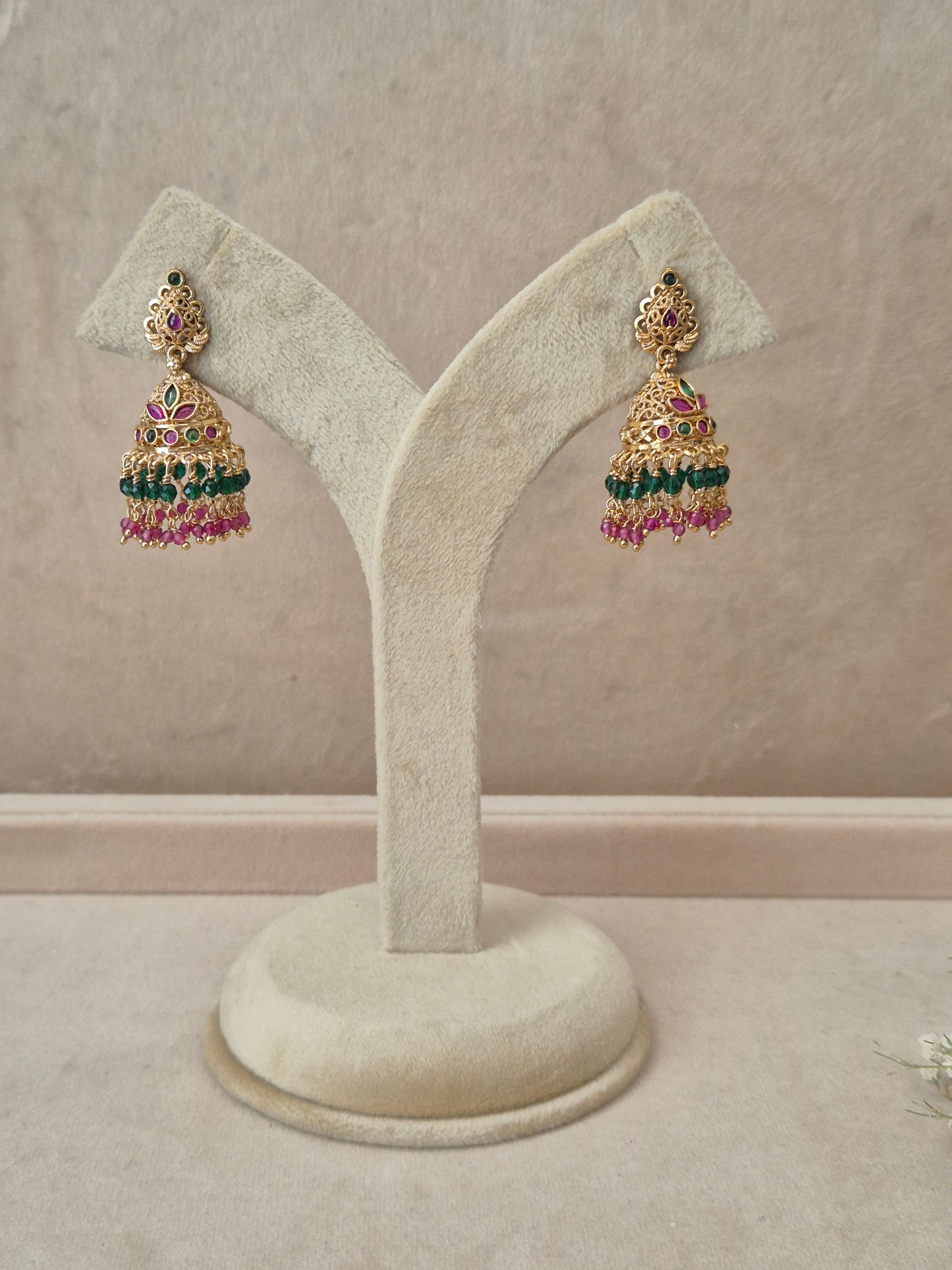 Pair of gold-plated Anamika Jhumka earrings hanging on a beige display stand, featuring traditional engraved studs with red and green gemstone accents and bell-shaped drops fringed with small green and pink beads by naitri.in