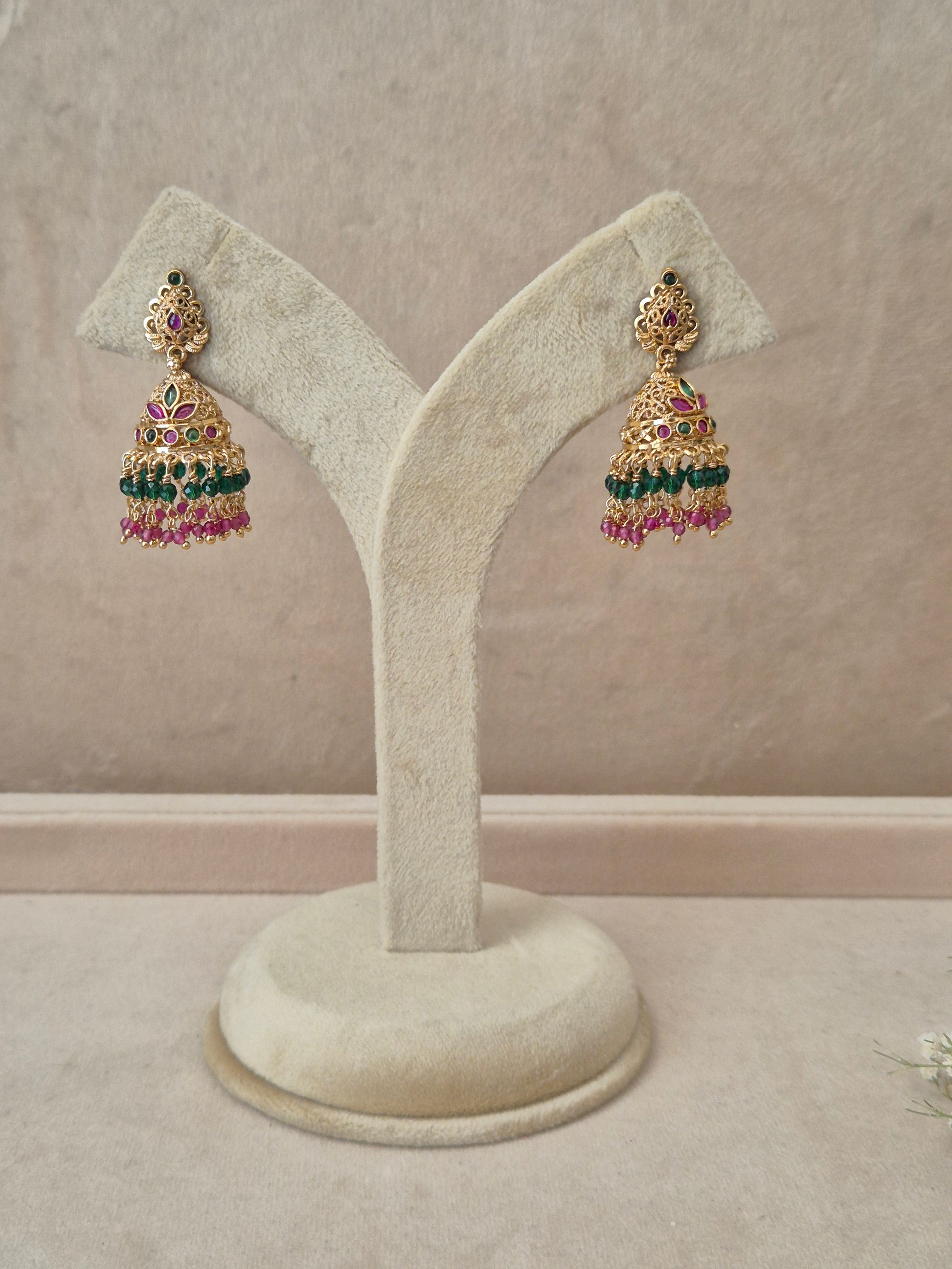 Pair of gold-plated Anamika Jhumka earrings hanging on a beige display stand, featuring traditional engraved studs with red and green gemstone accents and bell-shaped drops fringed with small green and pink beads by naitri.in