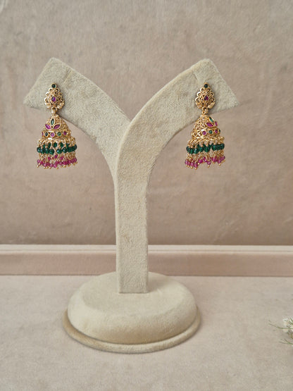 Pair of gold-plated Anamika Jhumka earrings hanging on a beige display stand, featuring traditional engraved studs with red and green gemstone accents and bell-shaped drops fringed with small green and pink beads by naitri.in