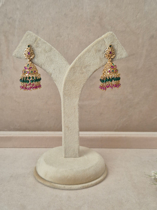 Pair of gold-plated Anamika Jhumka earrings hanging on a beige display stand, featuring traditional engraved studs with red and green gemstone accents and bell-shaped drops fringed with small green and pink beads by naitri.in