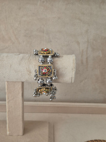 Dual-tone Apeksha bangle bracelet featuring a central square charm with a red and green floral enamel design and clear crystal center, flanked by oxidized silver beads and dangling metallic ghungroo balls by naitri.in
