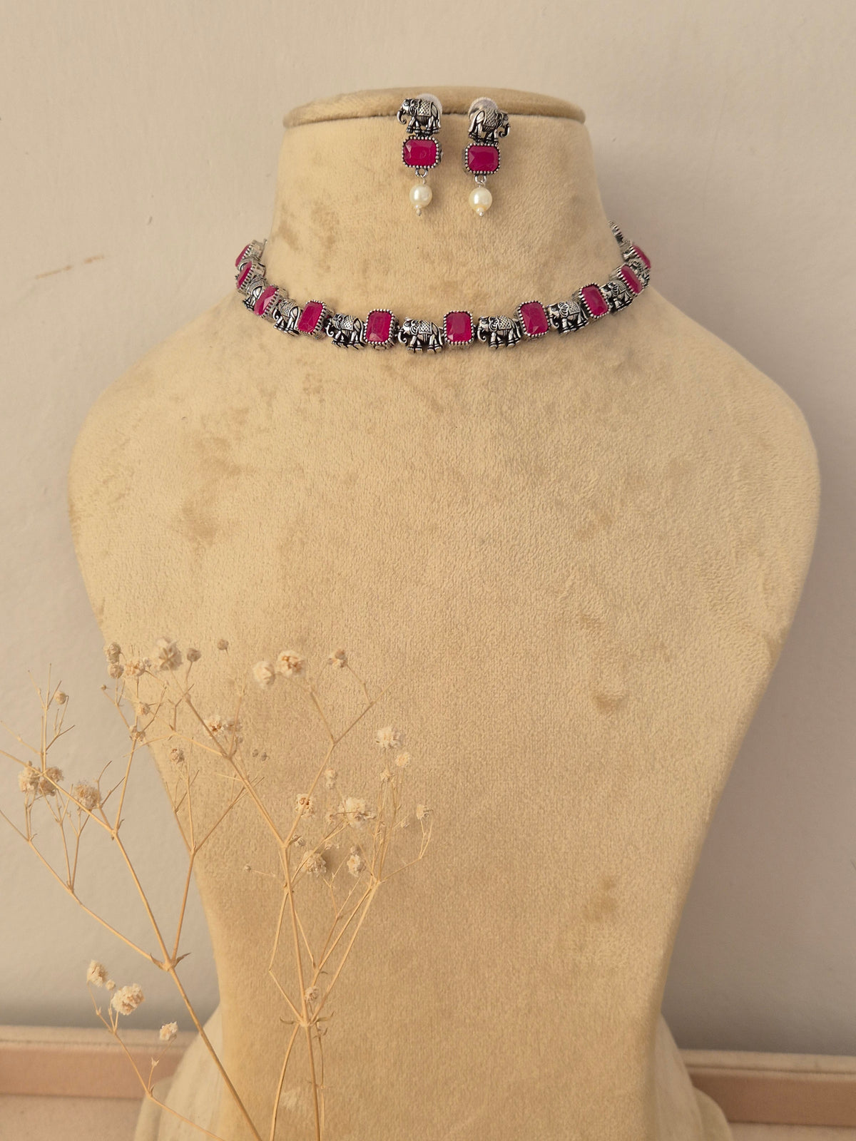 A jewelry set  featuring a silver-toned choker necklace with bright pink rectangular stones and matching drop earrings with pink stones and white pearl-like drops by naitri.in