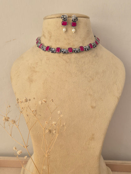 A jewelry set  featuring a silver-toned choker necklace with bright pink rectangular stones and matching drop earrings with pink stones and white pearl-like drops by naitri.in