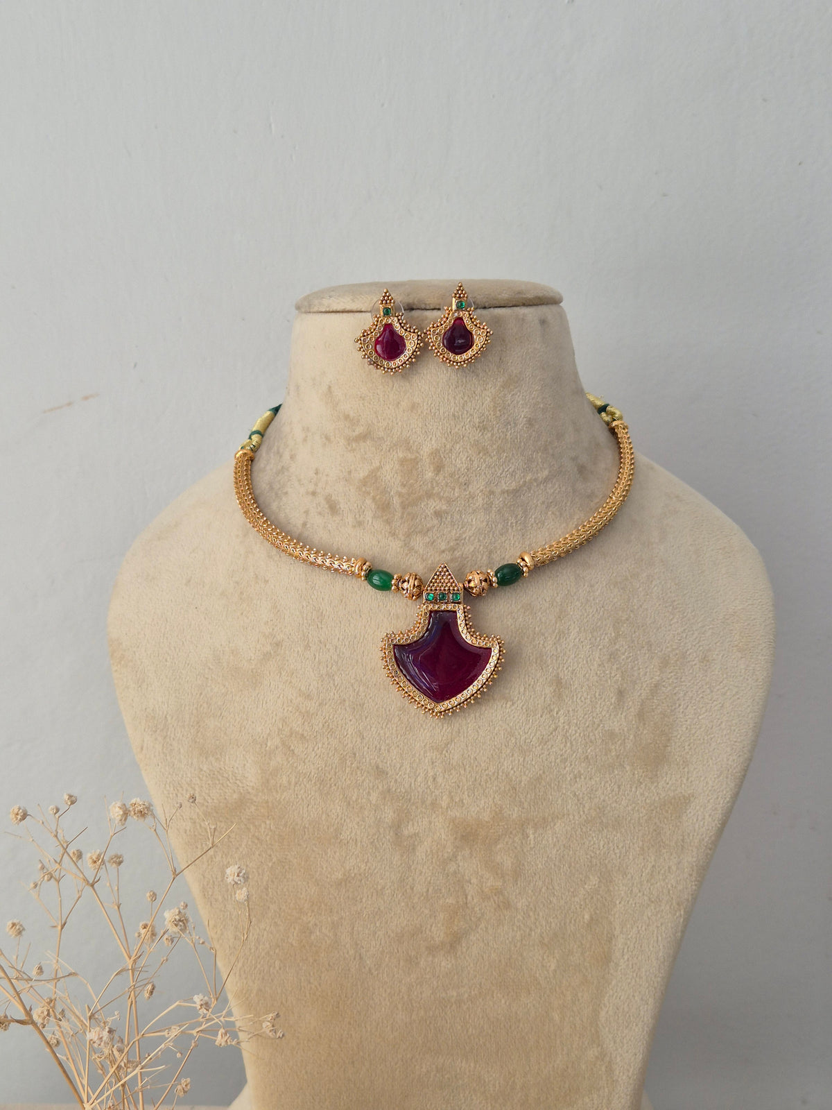 Astami gold-toned ethnic necklace set featuring a striking teardrop ruby-colored pendant, green beaded accents, and matching teardrop stud earrings by Naitri.in