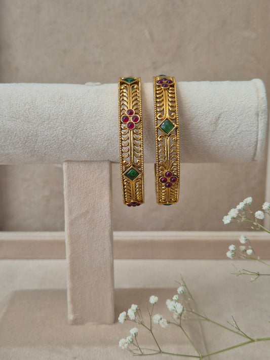 Pairs of gold Barkha bangles with intricate traditional carvings, featuring embedded red and green gemstones by naitri.in