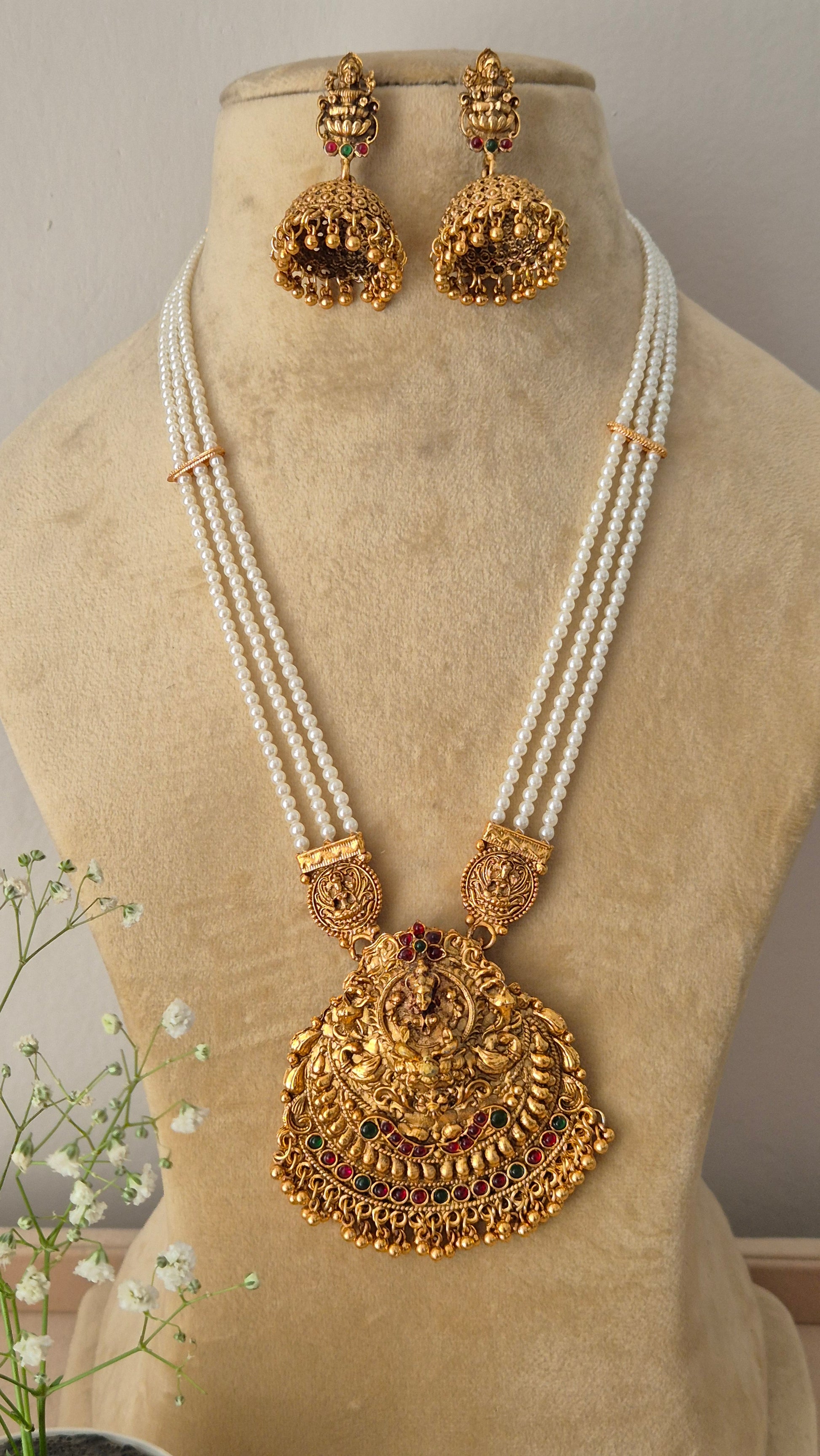 A traditional gold-plated temple jewelry pendant featuring an intricate goddess design, suspended from a multi-strand white pearl chain with  matching jhumka earrings by naitri.in