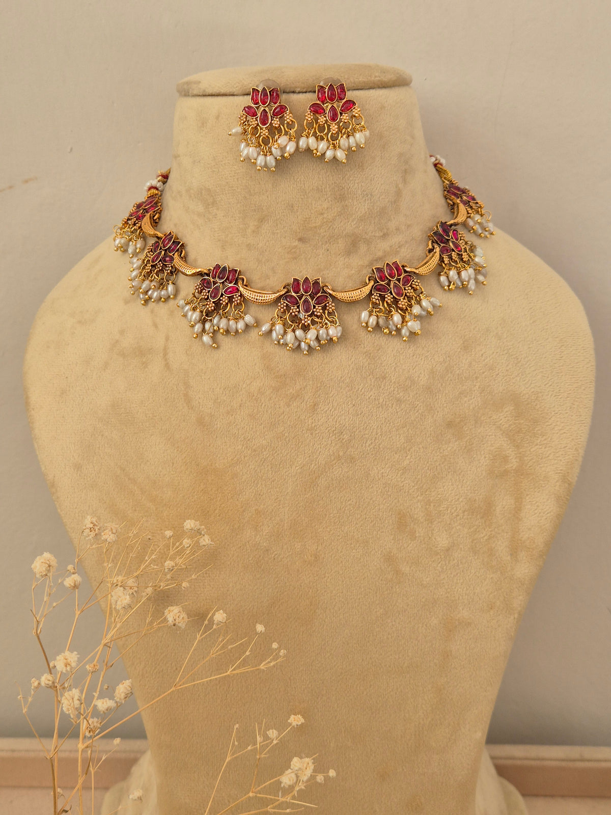 Gold-toned traditional Indian choker necklace and matching dangle earrings set features a series of delicate gold-toned floral motifs inset with vibrant red gemstones and a lower fringe of hanging small white pearls, by naitri.in