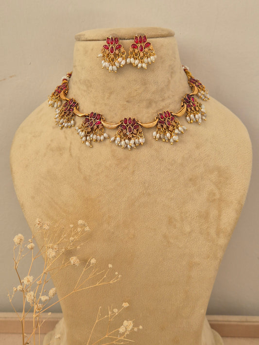 Gold-toned traditional Indian choker necklace and matching dangle earrings set features a series of delicate gold-toned floral motifs inset with vibrant red gemstones and a lower fringe of hanging small white pearls, by naitri.in