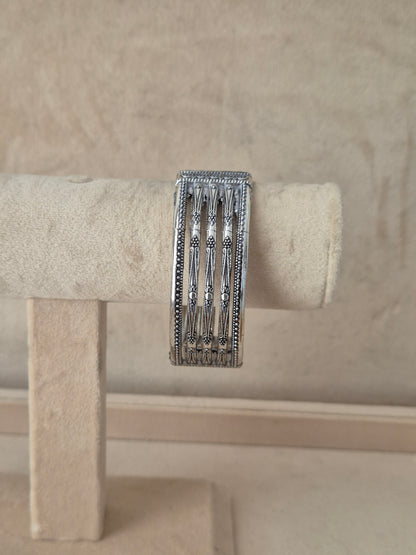 Oxidized silver Basanti bangle bracelet featuring a wide cuff design with vertical textured panels, open-work cutout slots, and small floral-like engravings, by naitri.in