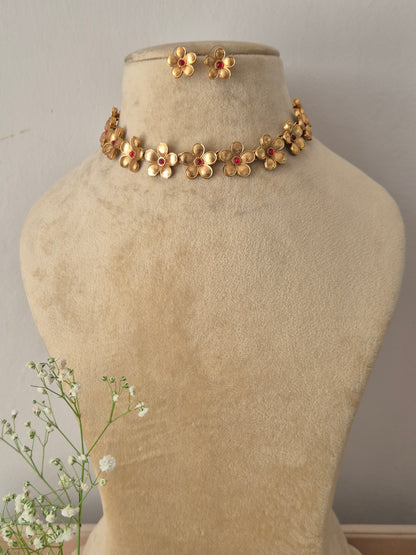 A gold-plated choker necklace featuring a row of intricate gold-toned flower charms with ruby-red gemstone centers, with matiching floral stud by naitri.in