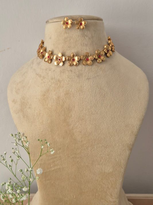 A gold-plated choker necklace featuring a row of intricate gold-toned flower charms with ruby-red gemstone centers, with matiching floral stud by naitri.in