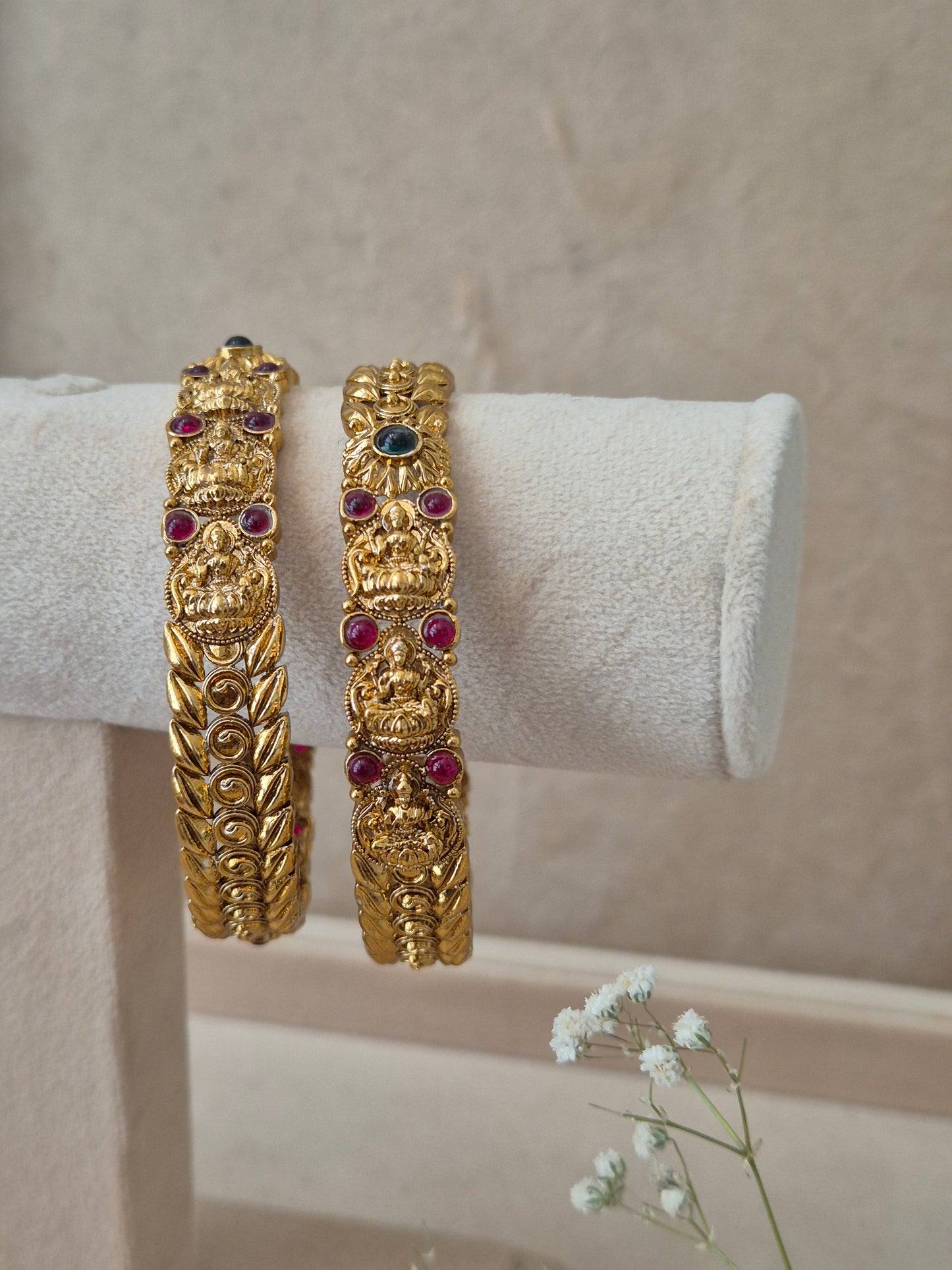 Pair of Naitri's Bhaghirathi gold bangles showcasing detailed ethnic craftsmanship, embedded with vibrant pinkish-red floral motifs and green accent stones,