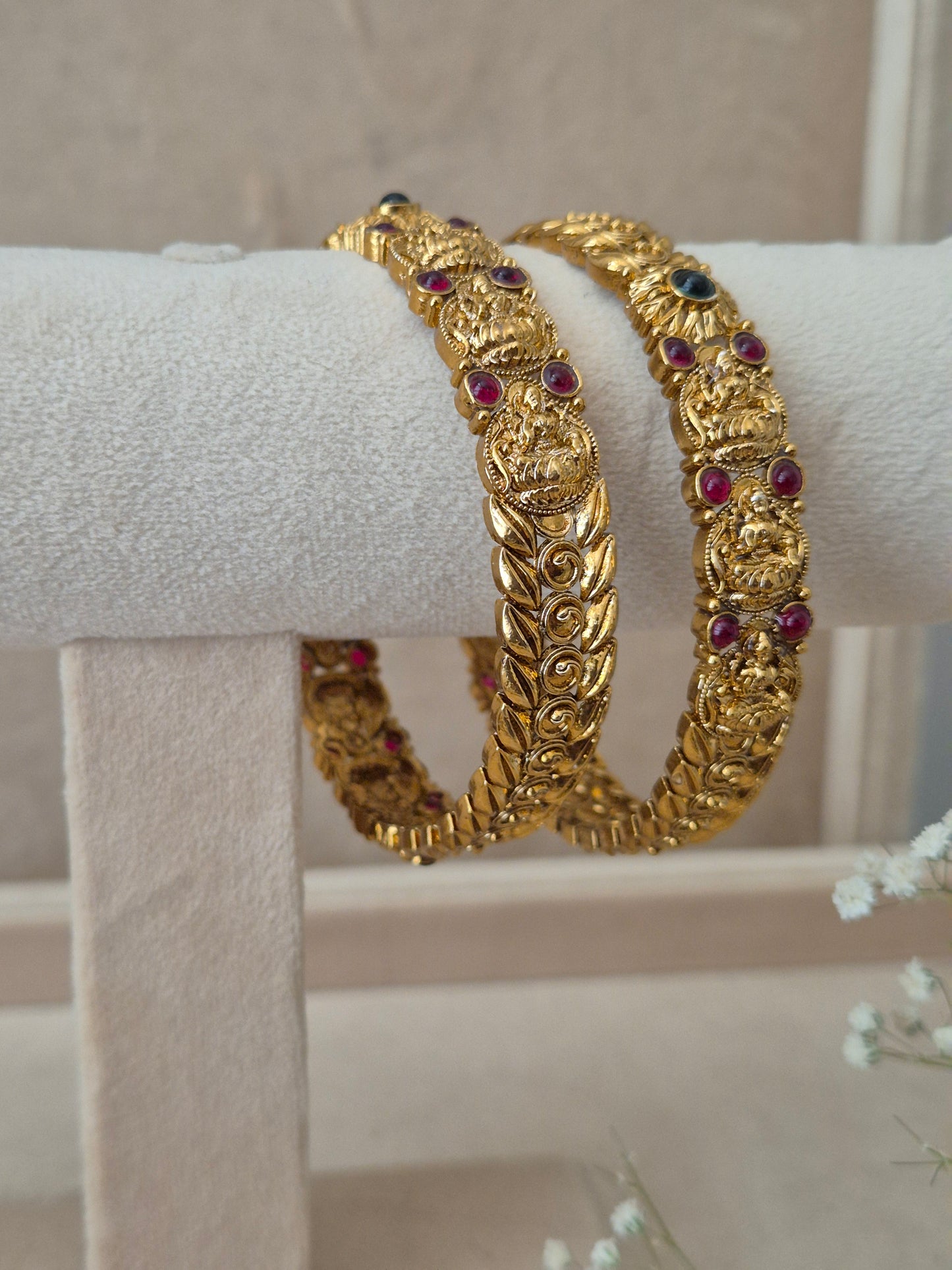 Two traditional gold-plated ethnic bangles with detailed leaf patterns and inlaid red and green gemstones on a white display stand by naitri.in