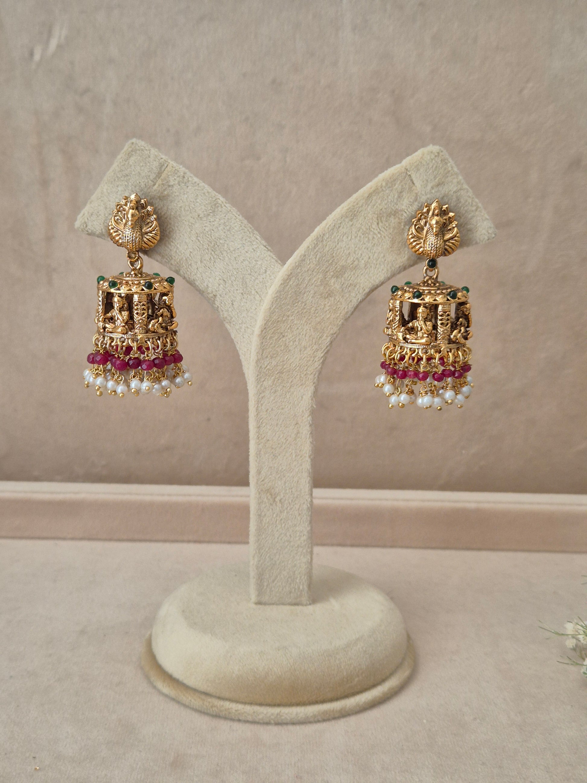 Pair of gold-plated Bhakti Jhumka earrings hanging on a beige velvet display stand, featuring detailed peacock motif studs, a band of green accents, and traditional bell-shaped drops lined with pink gemstones and white dangling pearls by naitri.in