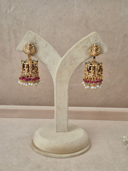 Pair of gold-plated Bhakti Jhumka earrings hanging on a beige velvet display stand, featuring detailed peacock motif studs, a band of green accents, and traditional bell-shaped drops lined with pink gemstones and white dangling pearls by naitri.in