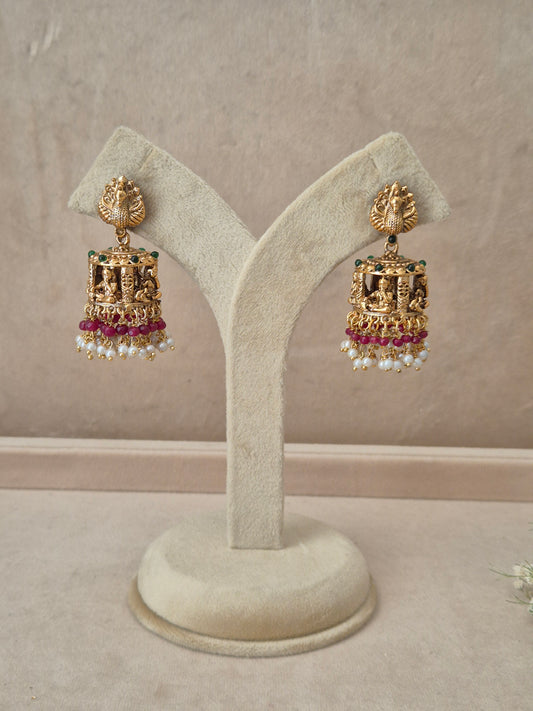 Pair of gold-plated Bhakti Jhumka earrings hanging on a beige velvet display stand, featuring detailed peacock motif studs, a band of green accents, and traditional bell-shaped drops lined with pink gemstones and white dangling pearls by naitri.in