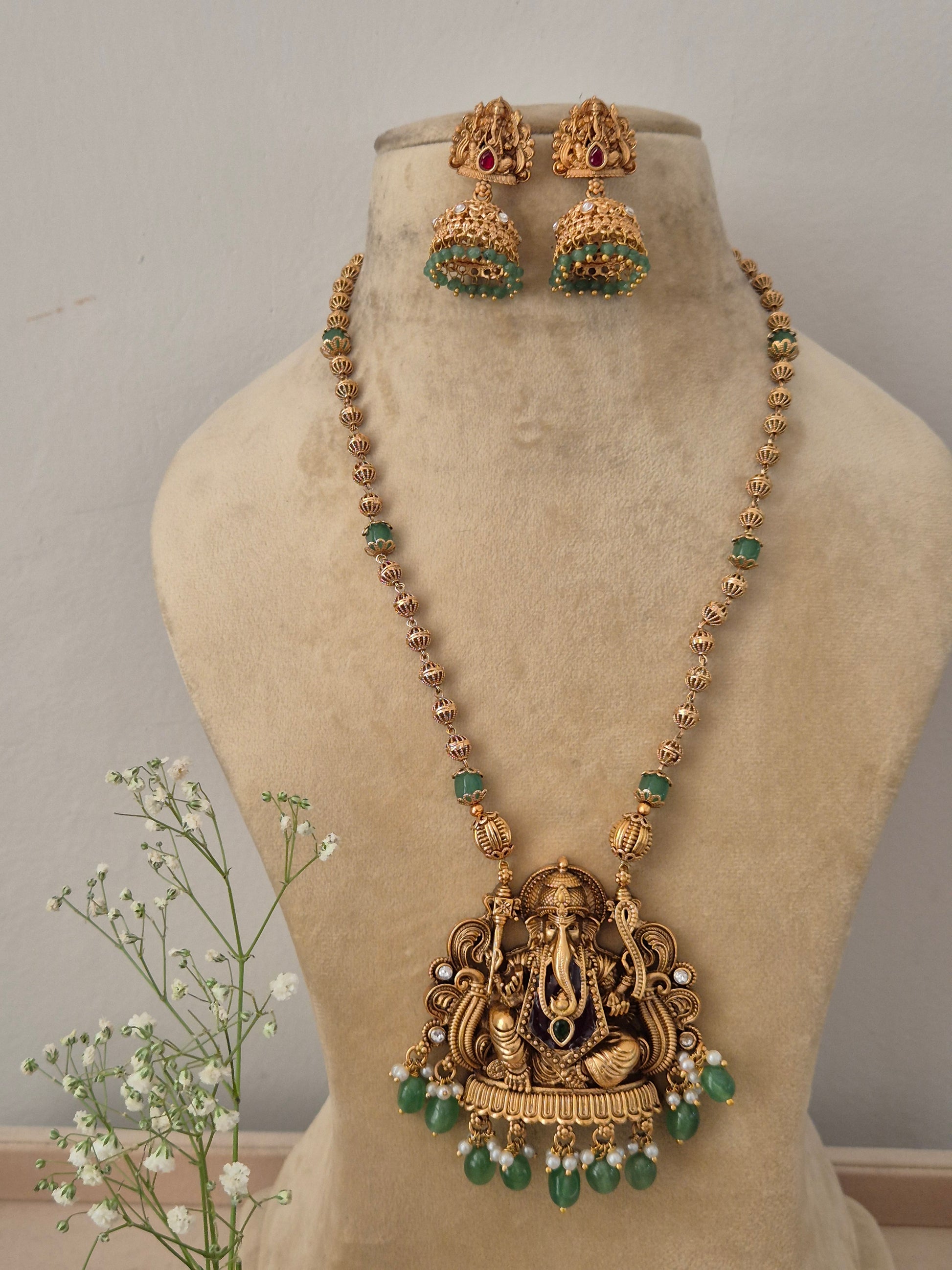 A gold-plated long necklace featuring an intricate Lord Ganesha temple pendant with dangling green beads with matching jhumka earrings by naitri.in