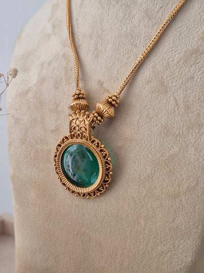 Traditional Indian gold-plated pendant necklace featuring a prominent round dark green gemstone bordered by tiny red stones, paired with matching round green stud earrings on a neutral mannequin by Naitri.in