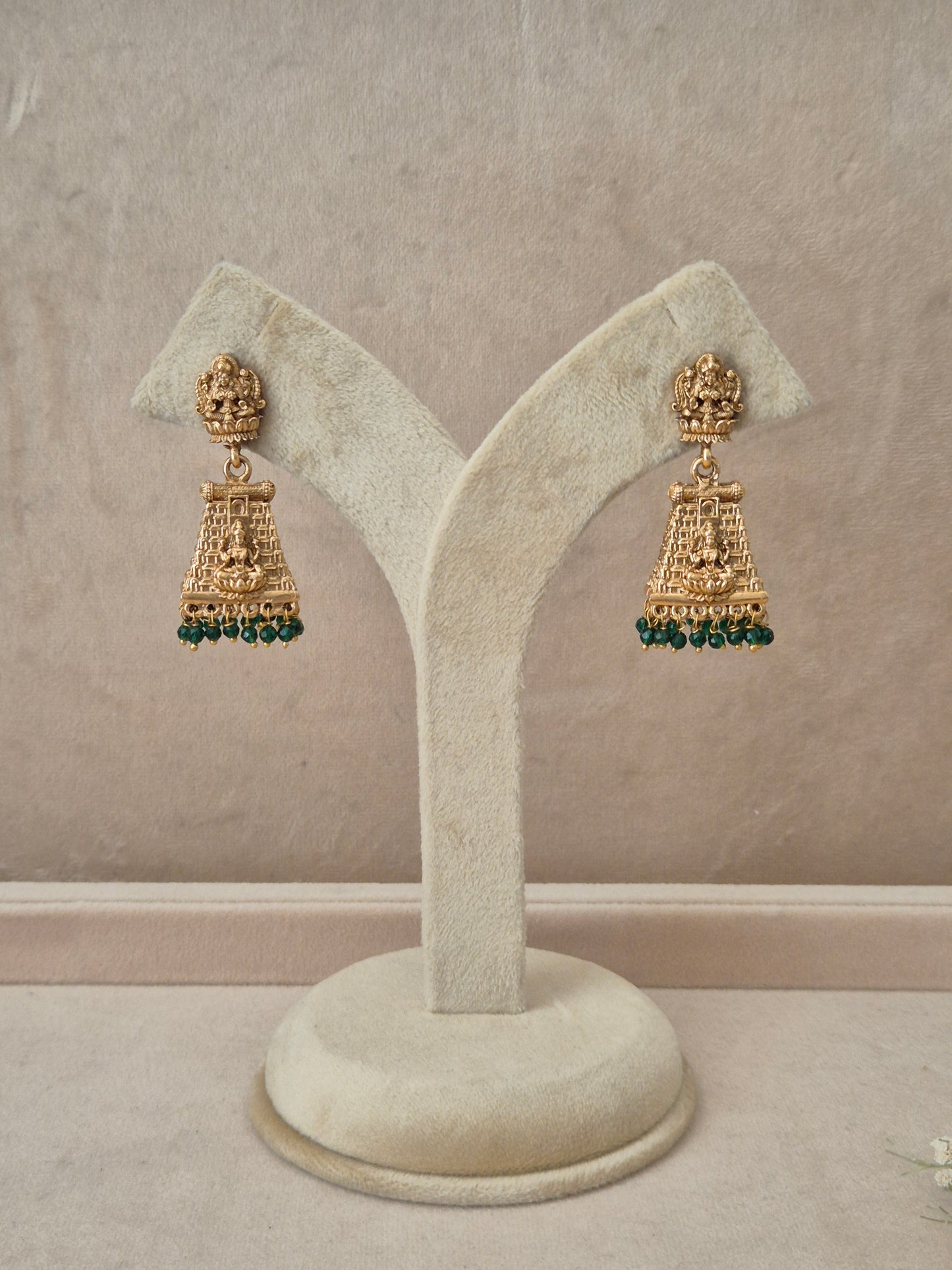 Pair of gold-toned Bhumika temple jewelry Jhumka earrings hanging on a beige velvet display stand, featuring rectangular Goddess motif studs and structured drops fringed with small green gemstone beads by naitri.in