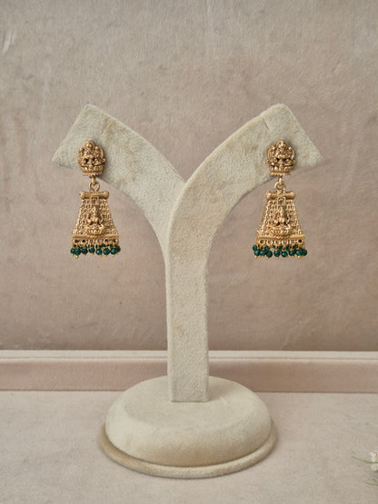 Pair of gold-toned Bhumika temple jewelry Jhumka earrings hanging on a beige velvet display stand, featuring rectangular Goddess motif studs and structured drops fringed with small green gemstone beads by naitri.in
