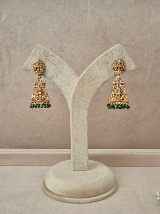 Pair of gold-toned Bhumika temple jewelry Jhumka earrings hanging on a beige velvet display stand, featuring rectangular Goddess motif studs and structured drops fringed with small green gemstone beads by naitri.in