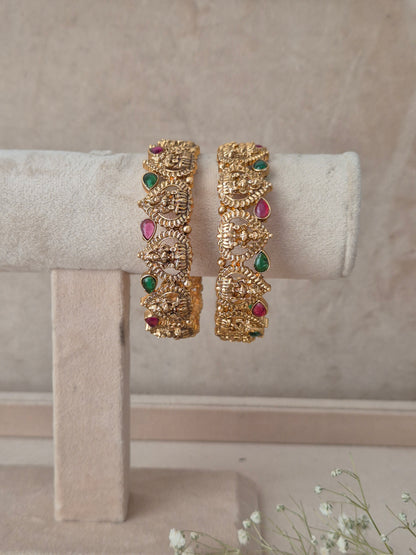 Festive antique gold bangle pair featuring carved temple motifs and colorful stone accents for wedding and traditional wear Antique gold-plated ethnic temple jewelry bangles with red and green stone detailing by naitri.in