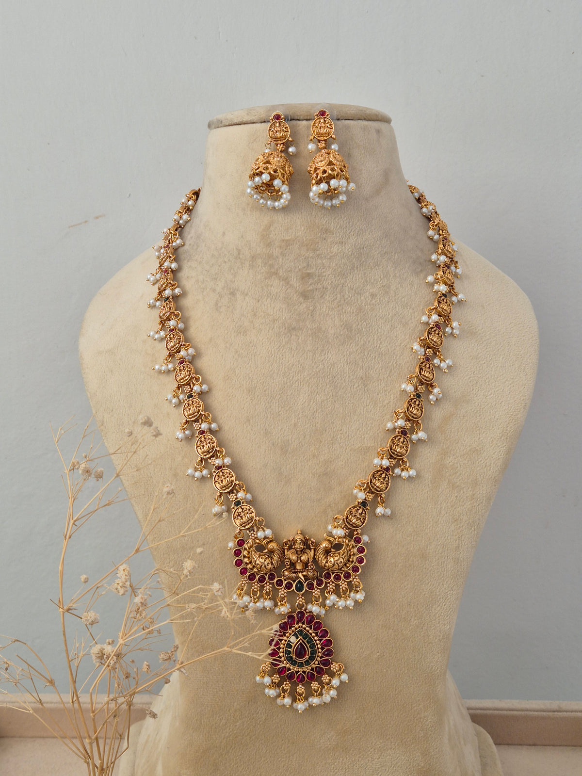 Bhuvneshwari Antique Gold Long Necklace Set displayed on a beige mannequin bust, showcasing intricate traditional temple craftsmanship with rich ruby stone accents and matching jhumka earrings by Naitri.in
