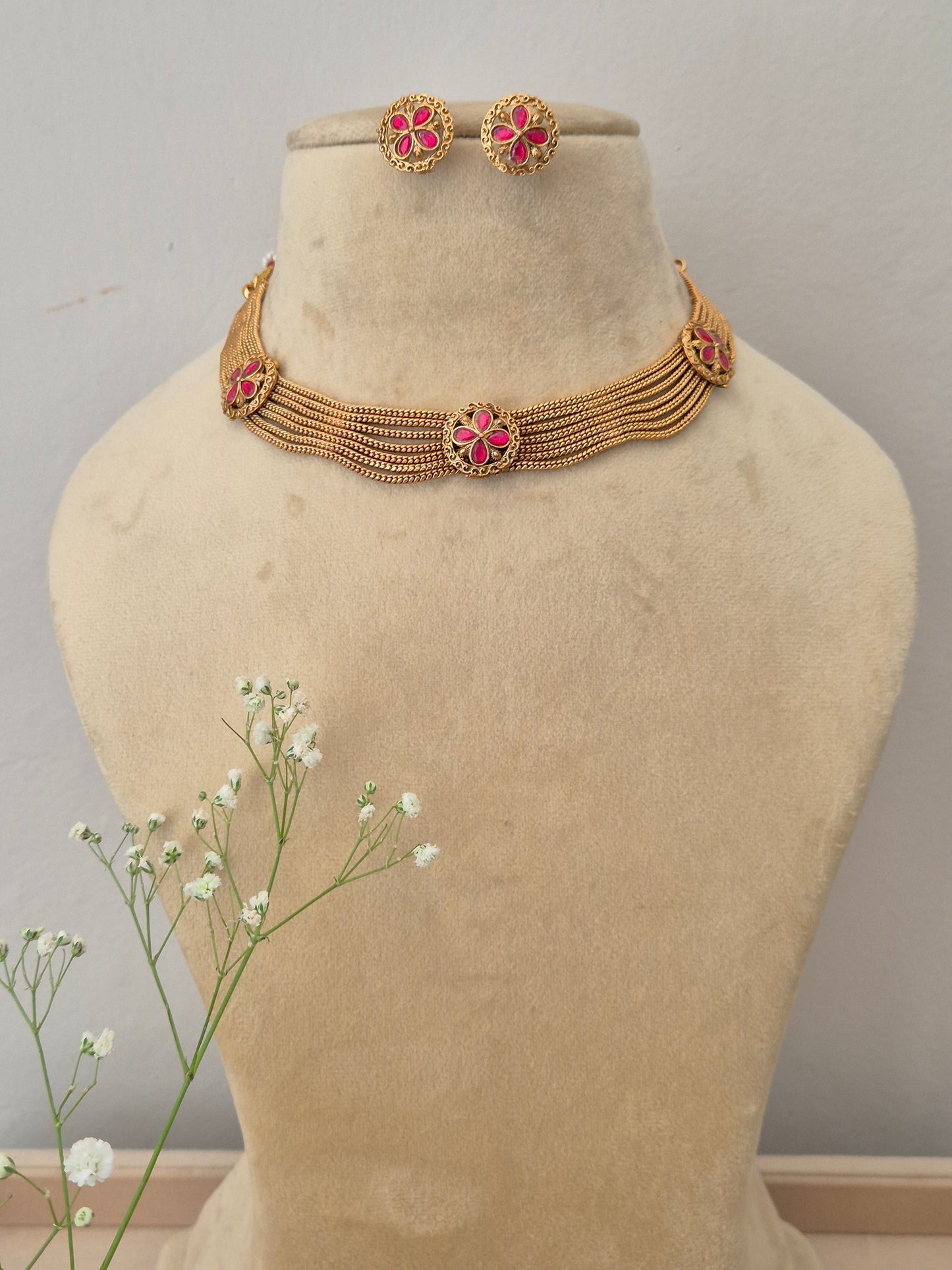 Bina Necklace Set A gold-plated multi-strand choker necklace featuring pink floral gemstone embellishments and matching stud earrings by naitri.in